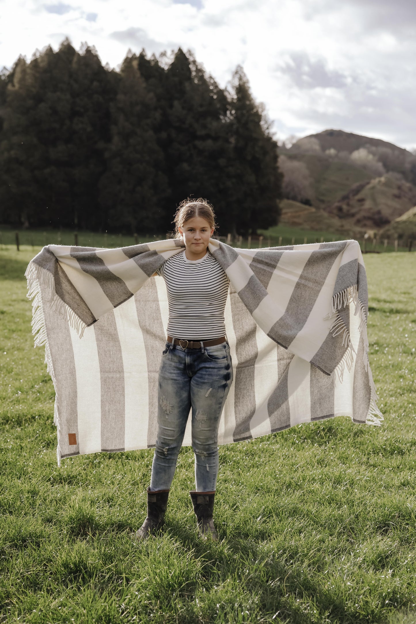 Person holding a large striped blanket in a grassy field with trees and mountains in the background