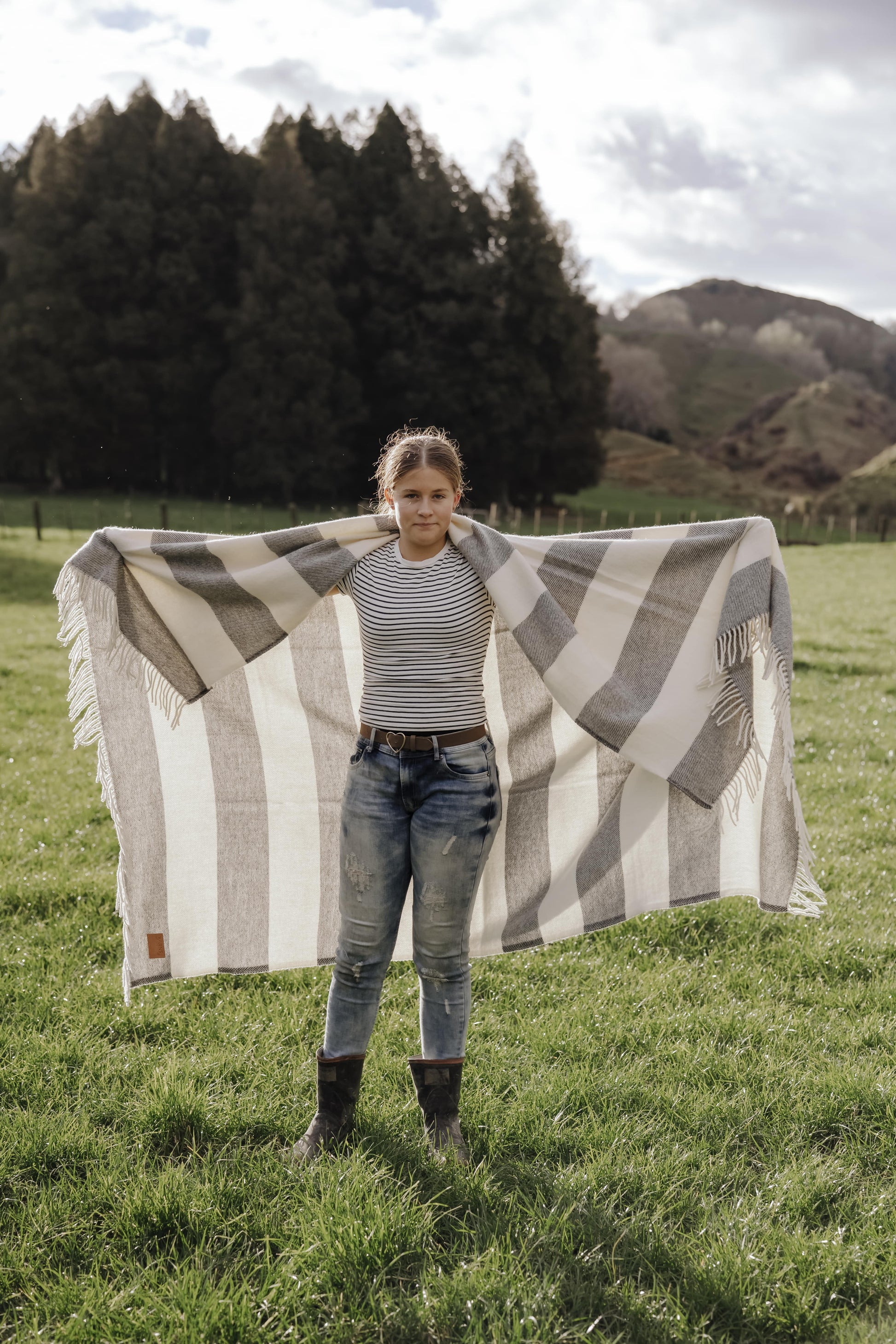 Person holding a large striped blanket in a grassy field with trees and mountains in the background