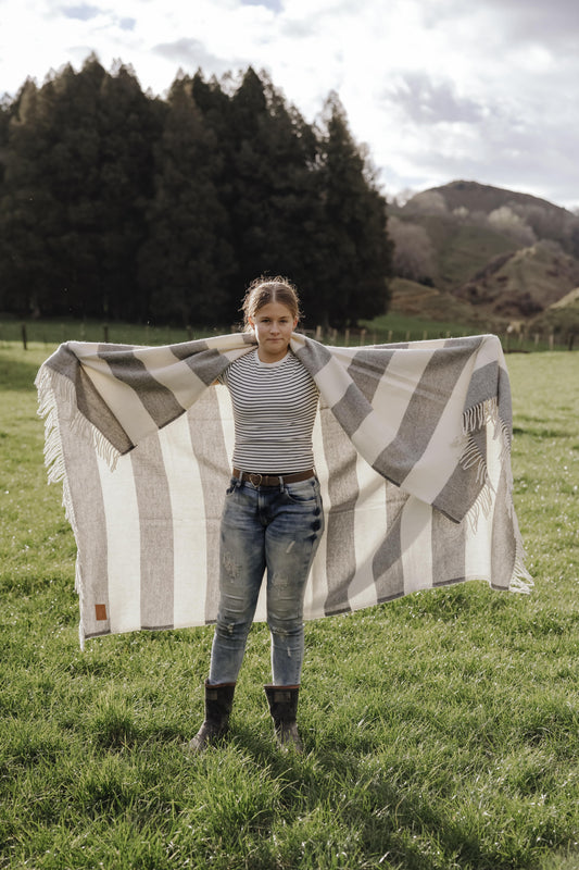Person holding a large striped blanket in a grassy field with trees and mountains in the background