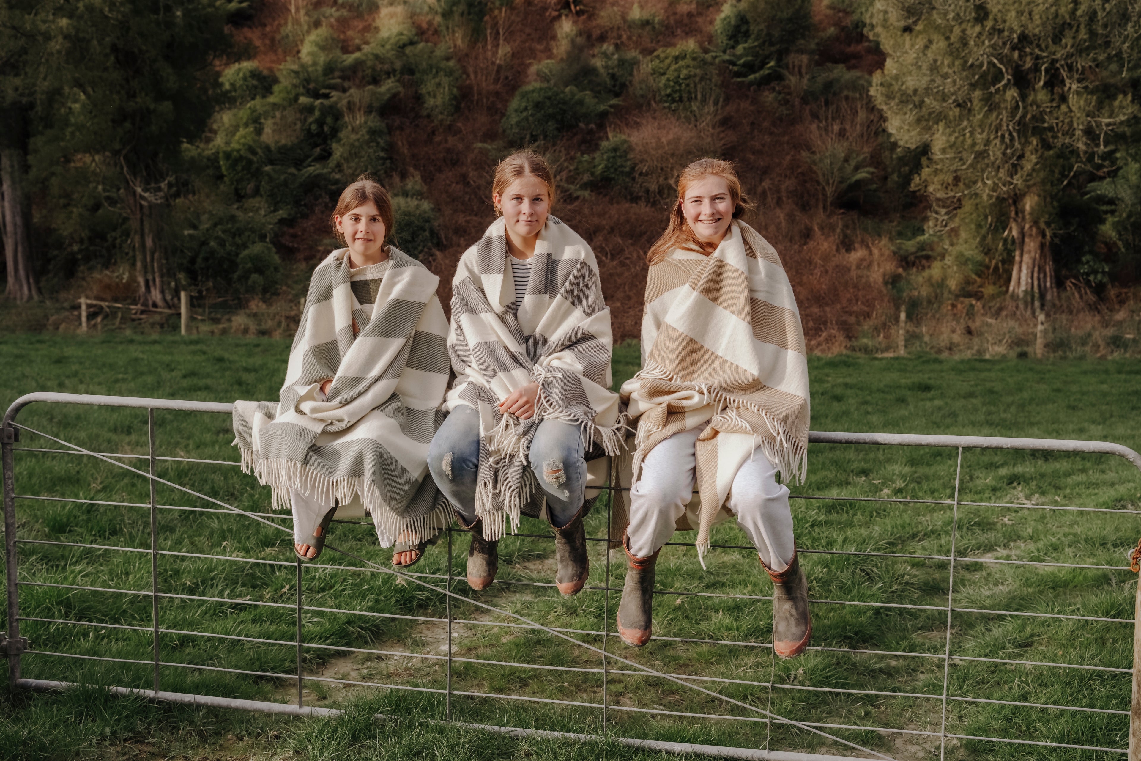 Three people sitting on a fence wearing patterned blankets in a natural setting.