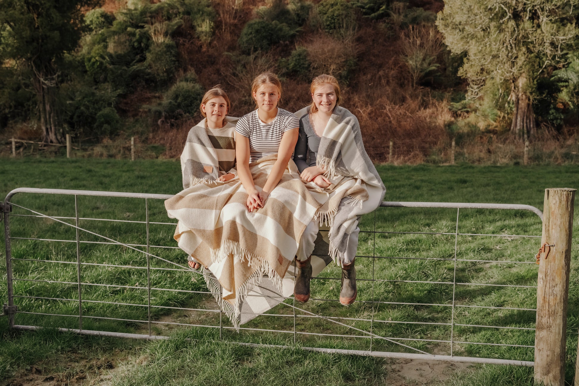 Three people sitting on a metal gate in a grassy field with trees in the background