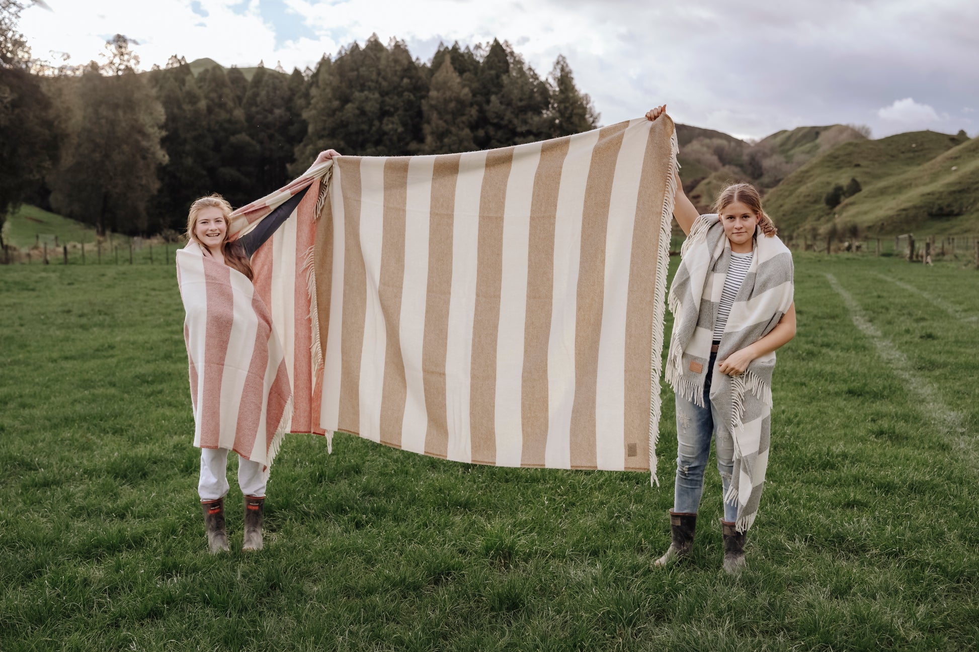 Two people holding a large striped blanket in a grassy field with trees and hills in the background.