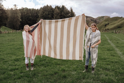 Two people holding a large striped blanket in a grassy field with trees and hills in the background.